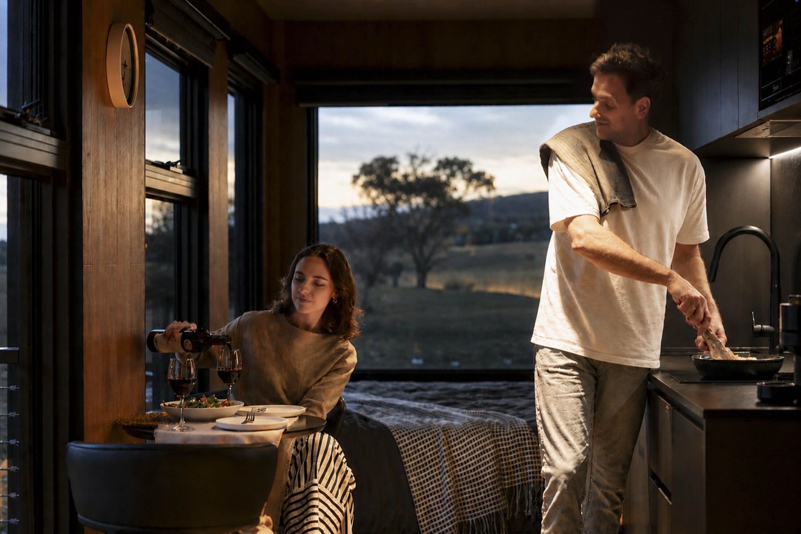 Guests cooking at the kitchen bench of an AUTRA retreat while wine is poured at the table, open country beyond the window